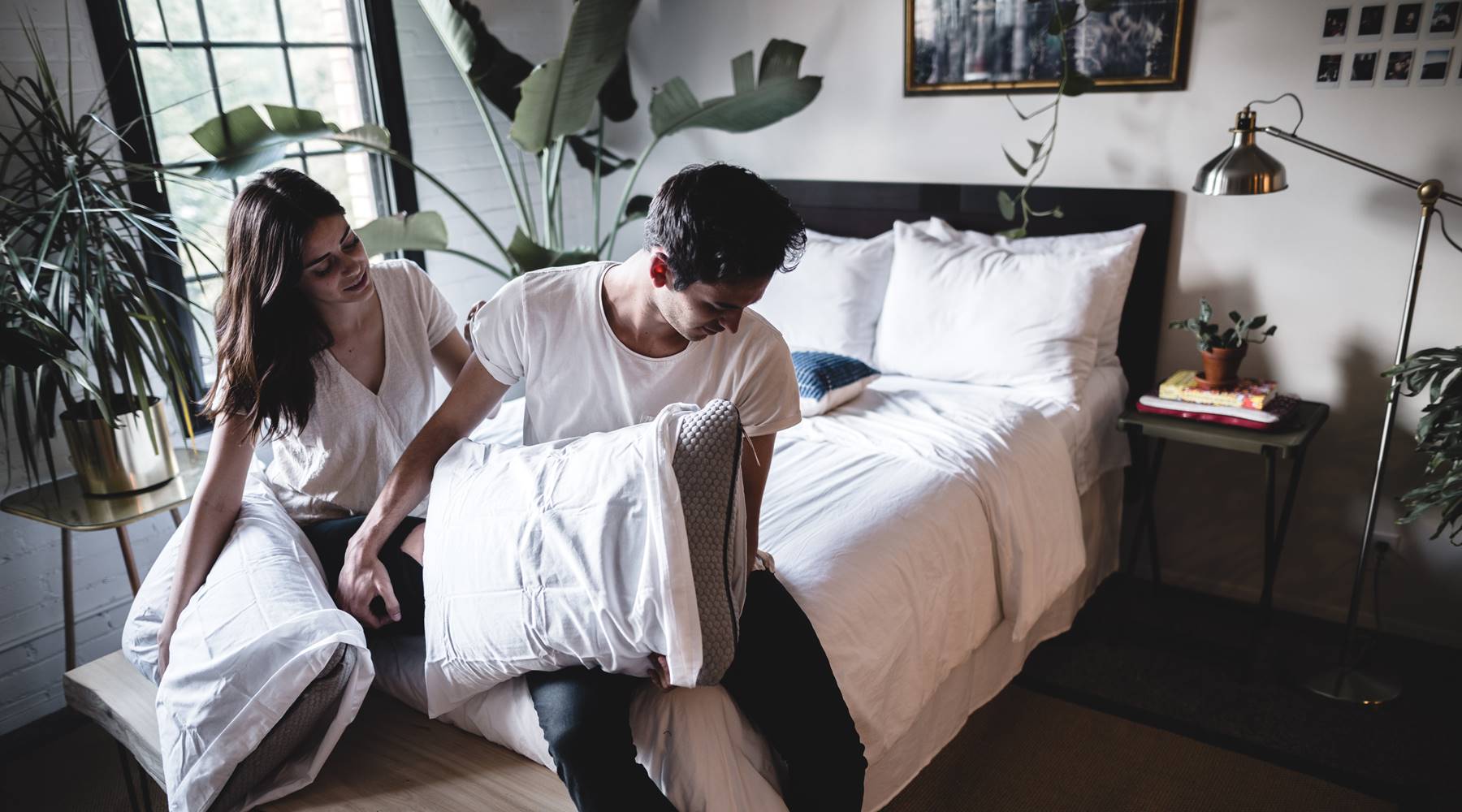 Man and Women in white t-shirts sitting on a bed holding pillows.